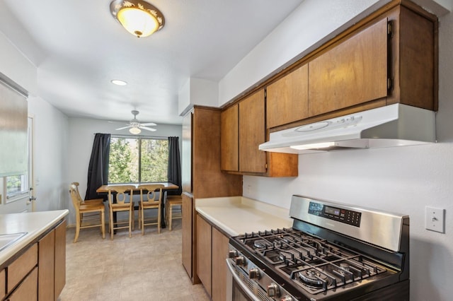 kitchen with stainless steel gas range, light countertops, under cabinet range hood, brown cabinets, and a ceiling fan
