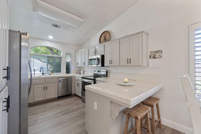 kitchen featuring vaulted ceiling, a sink, a peninsula, light wood finished floors, and stainless steel appliances