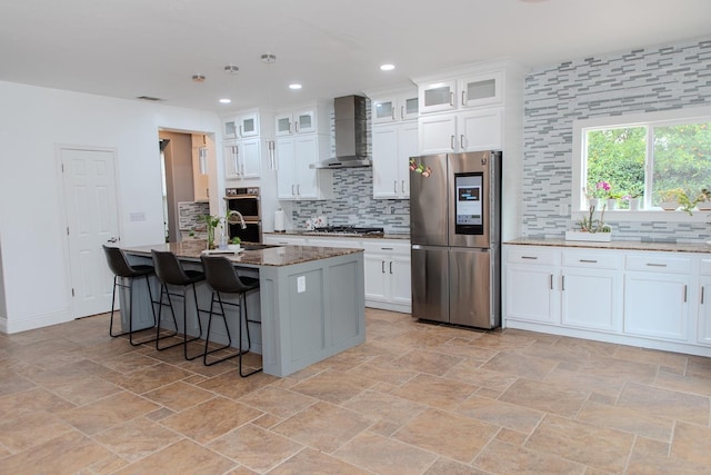 kitchen with wall chimney range hood, a kitchen island with sink, backsplash, appliances with stainless steel finishes, and white cabinets
