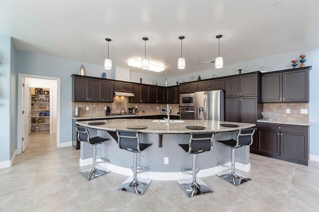 kitchen featuring dark brown cabinetry, backsplash, appliances with stainless steel finishes, under cabinet range hood, and light tile patterned flooring
