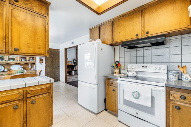 kitchen featuring brown cabinetry, white appliances, under cabinet range hood, tasteful backsplash, and light tile patterned floors