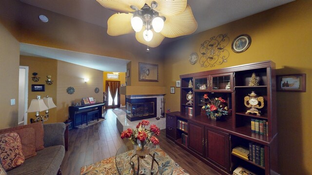 living area with a glass covered fireplace, ceiling fan, and dark wood-type flooring