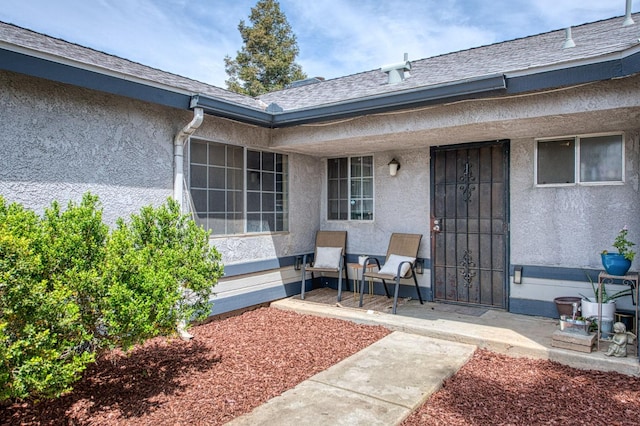 entrance to property with a shingled roof, stucco siding, and covered porch