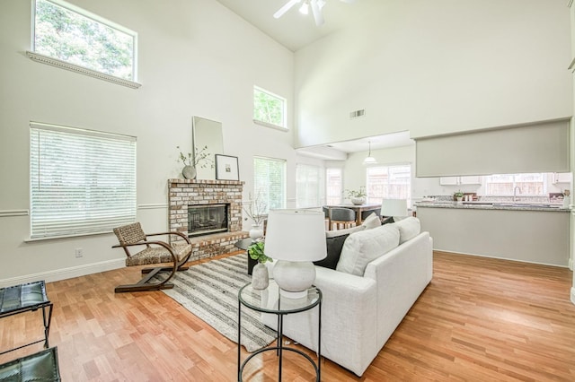 living room with a ceiling fan, visible vents, a towering ceiling, a brick fireplace, and light wood-style flooring