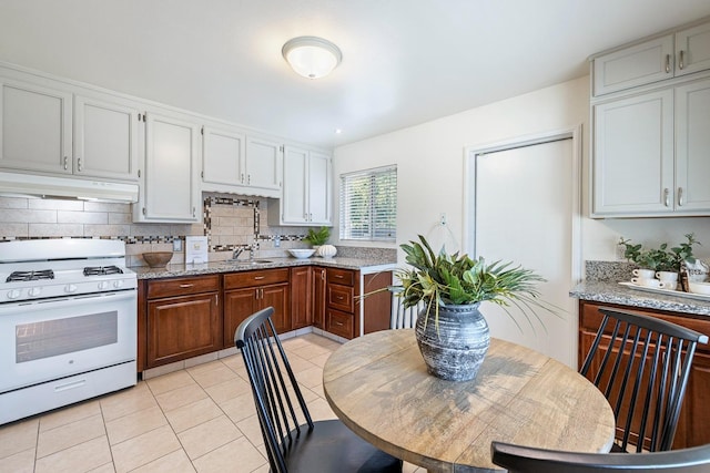 kitchen with white gas range oven, a sink, decorative backsplash, light tile patterned floors, and under cabinet range hood