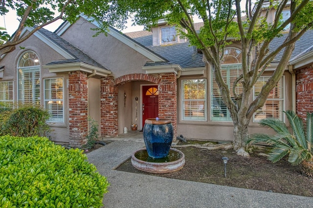 view of exterior entry featuring brick siding, a shingled roof, and stucco siding