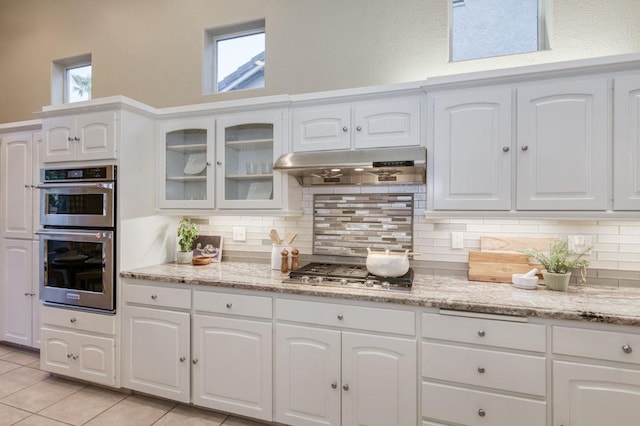 kitchen with under cabinet range hood, backsplash, appliances with stainless steel finishes, and glass insert cabinets