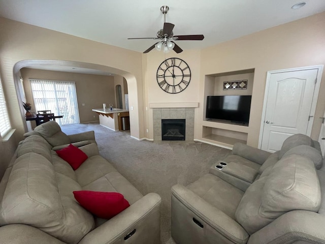 carpeted living area featuring built in features, baseboards, ceiling fan, a tiled fireplace, and arched walkways