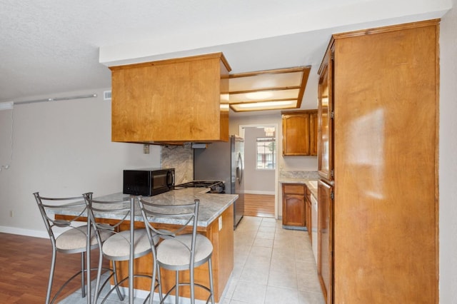 kitchen featuring brown cabinetry, freestanding refrigerator, black microwave, a breakfast bar, and light countertops