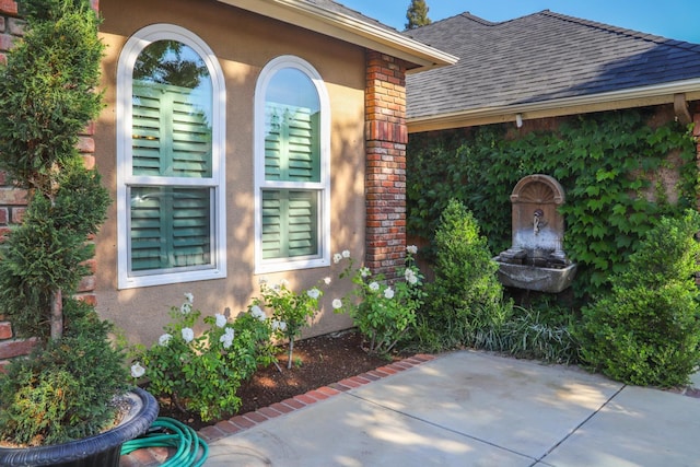 view of exterior entry with brick siding, a shingled roof, a patio area, and stucco siding