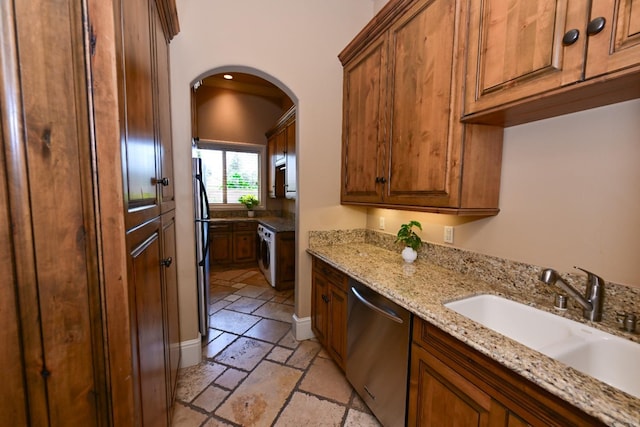 kitchen with brown cabinets, a sink, arched walkways, stone tile floors, and stainless steel dishwasher