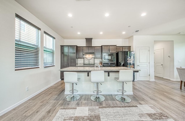 kitchen featuring a breakfast bar, wall oven, an island with sink, built in microwave, and light wood-style flooring
