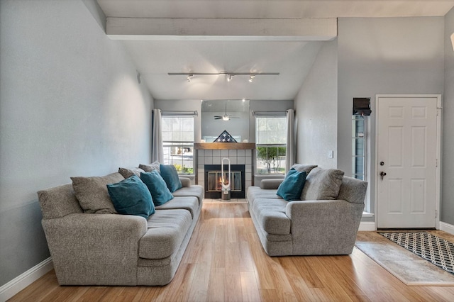 living area with lofted ceiling with beams, light wood finished floors, rail lighting, a tiled fireplace, and baseboards