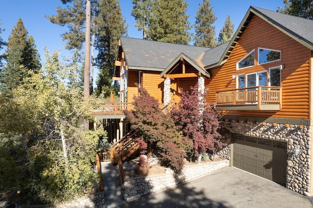 log-style house with stone siding, an attached garage, a shingled roof, a balcony, and driveway