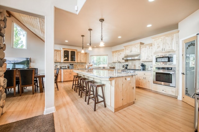kitchen with light wood finished floors, glass insert cabinets, a kitchen breakfast bar, a kitchen island with sink, and appliances with stainless steel finishes