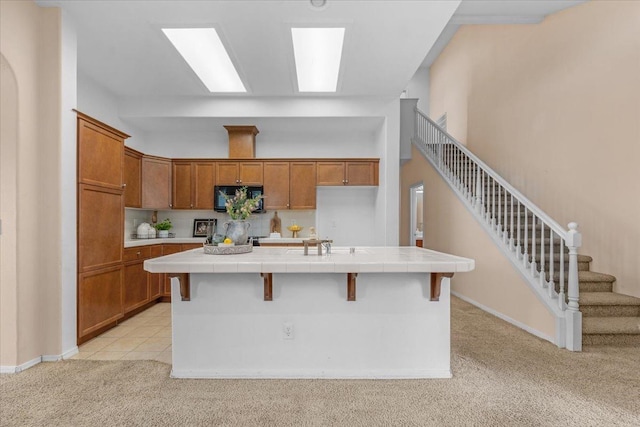 kitchen featuring light colored carpet, brown cabinetry, black microwave, and tile counters