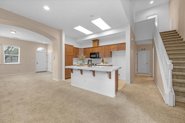 kitchen with brown cabinets, an island with sink, arched walkways, light carpet, and black microwave