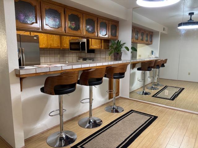 7093 N Teilman Unit 102, Fresno, CA 93711 - kitchen featuring freestanding refrigerator, brown cabinetry, stainless steel microwave, light wood-type flooring, and a breakfast bar area