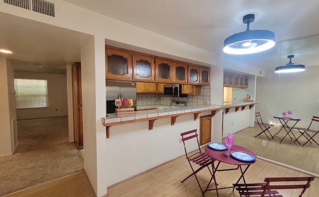 kitchen featuring brown cabinetry, stainless steel appliances, tile countertops, light wood finished floors, and glass insert cabinets