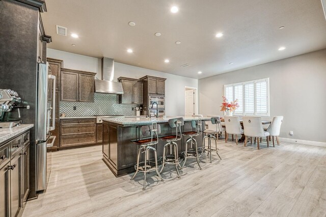 kitchen featuring wall chimney exhaust hood, a kitchen breakfast bar, a center island with sink, visible vents, and light wood-style floors