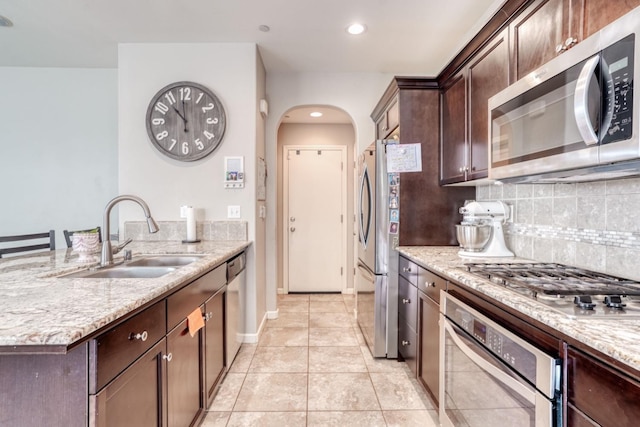 kitchen with a sink, backsplash, arched walkways, dark brown cabinets, and appliances with stainless steel finishes