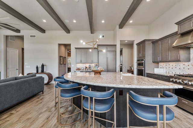 kitchen featuring stainless steel appliances, decorative backsplash, beam ceiling, wall chimney range hood, and light wood finished floors