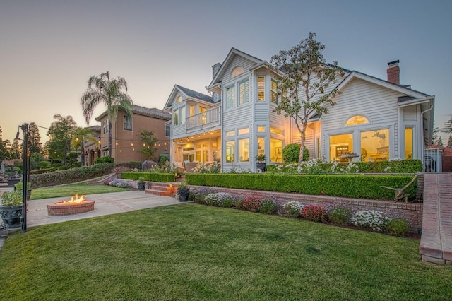 back of house at dusk featuring an outdoor fire pit, a balcony, a patio area, and a lawn