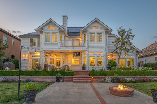 back of house at dusk with a patio area, a balcony, solar panels, a lawn, and an outdoor fire pit