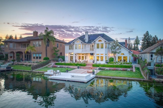 back of house at dusk with a water view, a chimney, and a balcony
