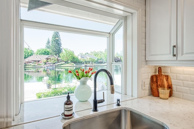 interior details with light stone counters, a sink, white cabinetry, a water view, and decorative backsplash
