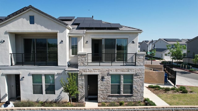 view of front facade featuring a balcony, stucco siding, roof mounted solar panels, and stone siding