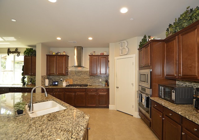 kitchen featuring wall chimney range hood, a sink, appliances with stainless steel finishes, backsplash, and light stone counters