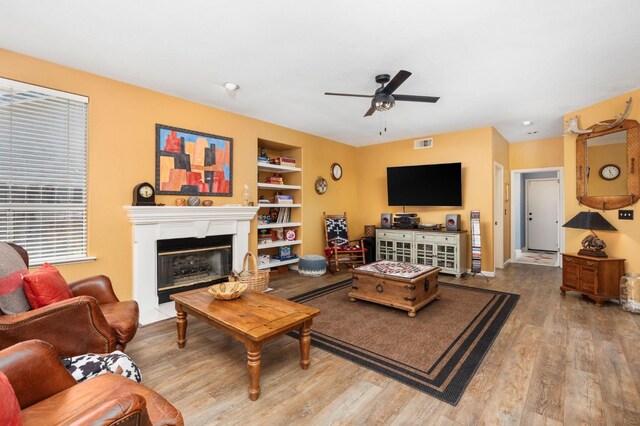 living room featuring a ceiling fan, built in features, wood finished floors, and a glass covered fireplace