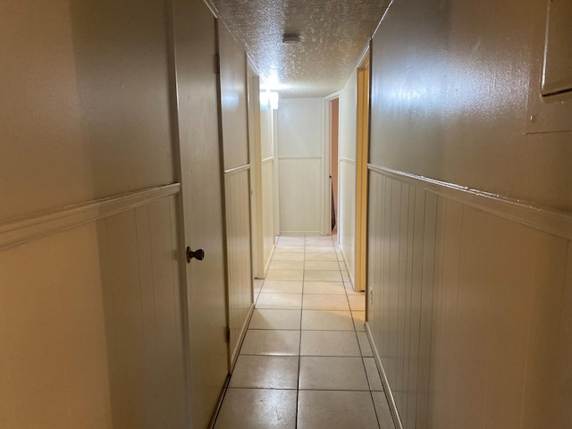 hallway with a textured ceiling, light tile patterned floors, a decorative wall, and wainscoting