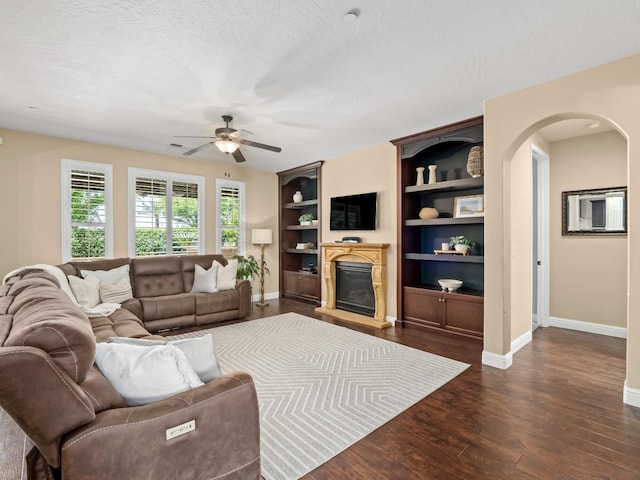 living area featuring baseboards, ceiling fan, a textured ceiling, a glass covered fireplace, and dark wood-type flooring
