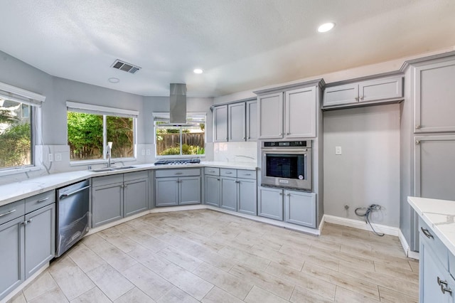 kitchen with appliances with stainless steel finishes, gray cabinets, island range hood, a sink, and light stone counters