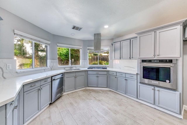 kitchen with stainless steel appliances, gray cabinets, a sink, island exhaust hood, and backsplash