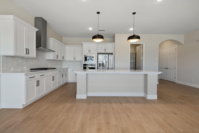 kitchen with appliances with stainless steel finishes, wall chimney exhaust hood, tasteful backsplash, white cabinets, and light wood-type flooring