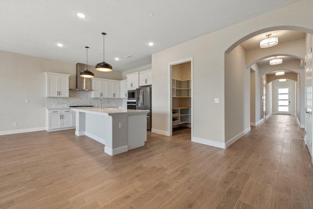 kitchen with stainless steel appliances, wall chimney exhaust hood, light countertops, white cabinets, and a center island with sink
