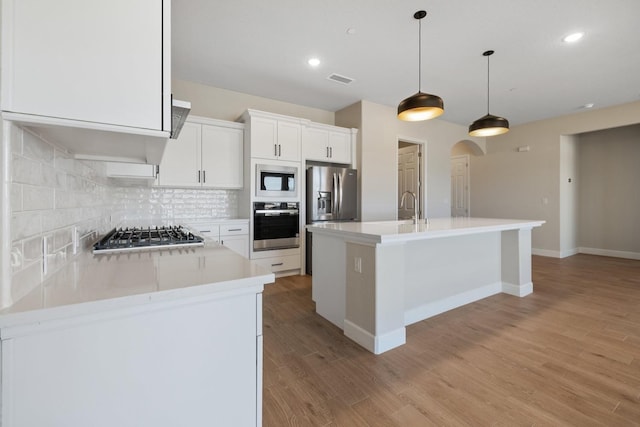 kitchen with stainless steel appliances, an island with sink, decorative backsplash, light wood-type flooring, and arched walkways