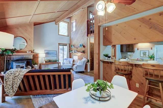 dining room featuring wood walls, a ceiling fan, wood finished floors, a tiled fireplace, and high vaulted ceiling