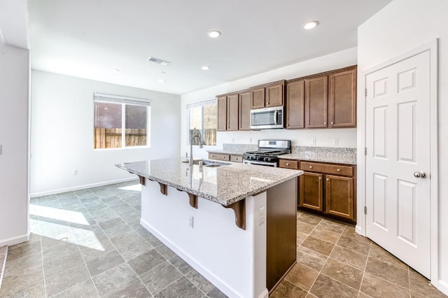 kitchen with stainless steel appliances, a sink, a kitchen island with sink, light stone countertops, and a kitchen bar