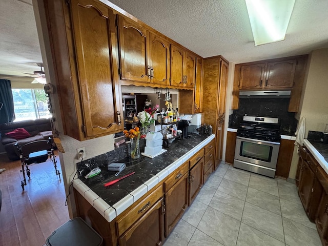 kitchen featuring gas stove, under cabinet range hood, tile counters, brown cabinetry, and a textured ceiling