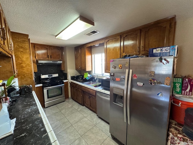 kitchen featuring appliances with stainless steel finishes, under cabinet range hood, brown cabinets, tile countertops, and a textured ceiling