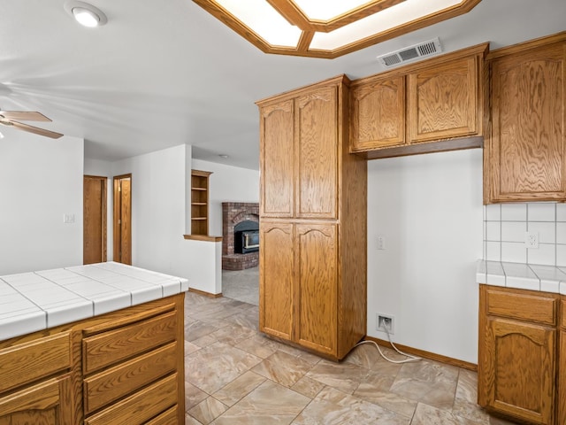 kitchen featuring a ceiling fan, tile countertops, a brick fireplace, backsplash, and brown cabinetry