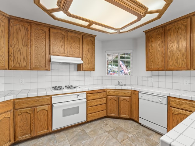 kitchen featuring white appliances, under cabinet range hood, brown cabinetry, and light countertops
