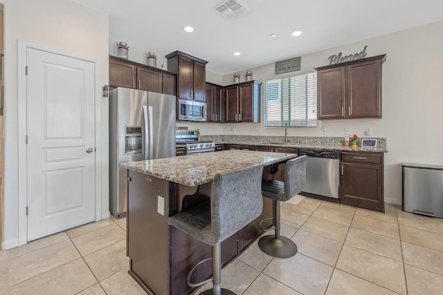 kitchen featuring appliances with stainless steel finishes, a sink, dark brown cabinets, a breakfast bar, and light stone counters