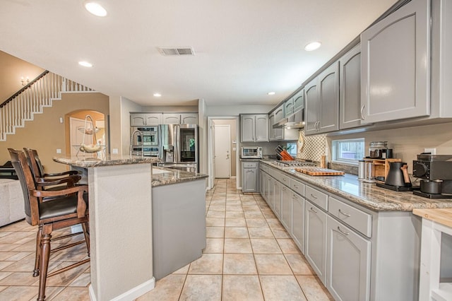 kitchen featuring gray cabinets, appliances with stainless steel finishes, light stone countertops, a kitchen bar, and light tile patterned floors