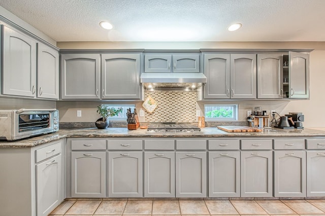 kitchen with gray cabinets, a textured ceiling, stainless steel gas stovetop, under cabinet range hood, and light tile patterned floors