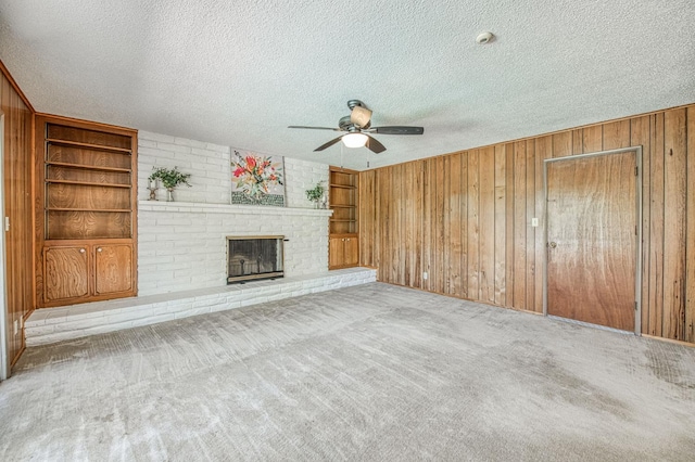 unfurnished living room featuring a ceiling fan, wooden walls, a textured ceiling, built in shelves, and a brick fireplace